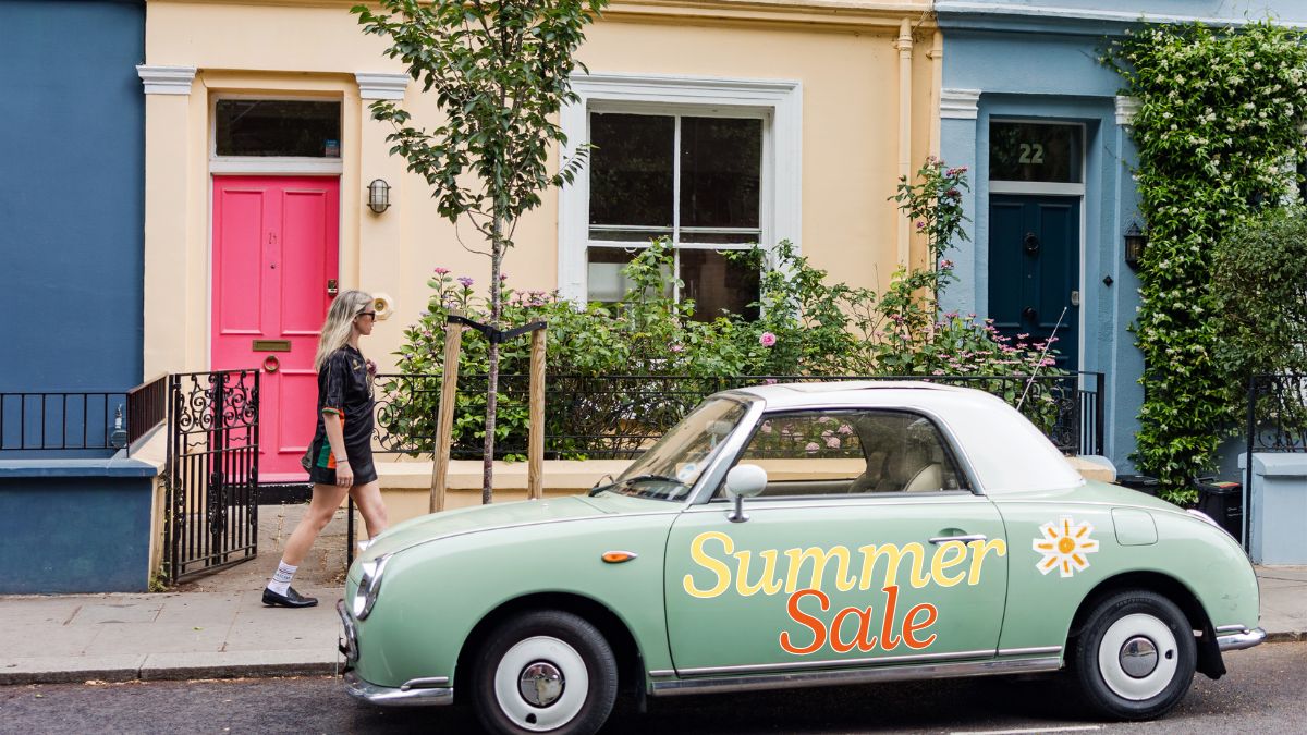 Vintage green car with 'Summer Sale' text parked on a street in front of colorful houses.