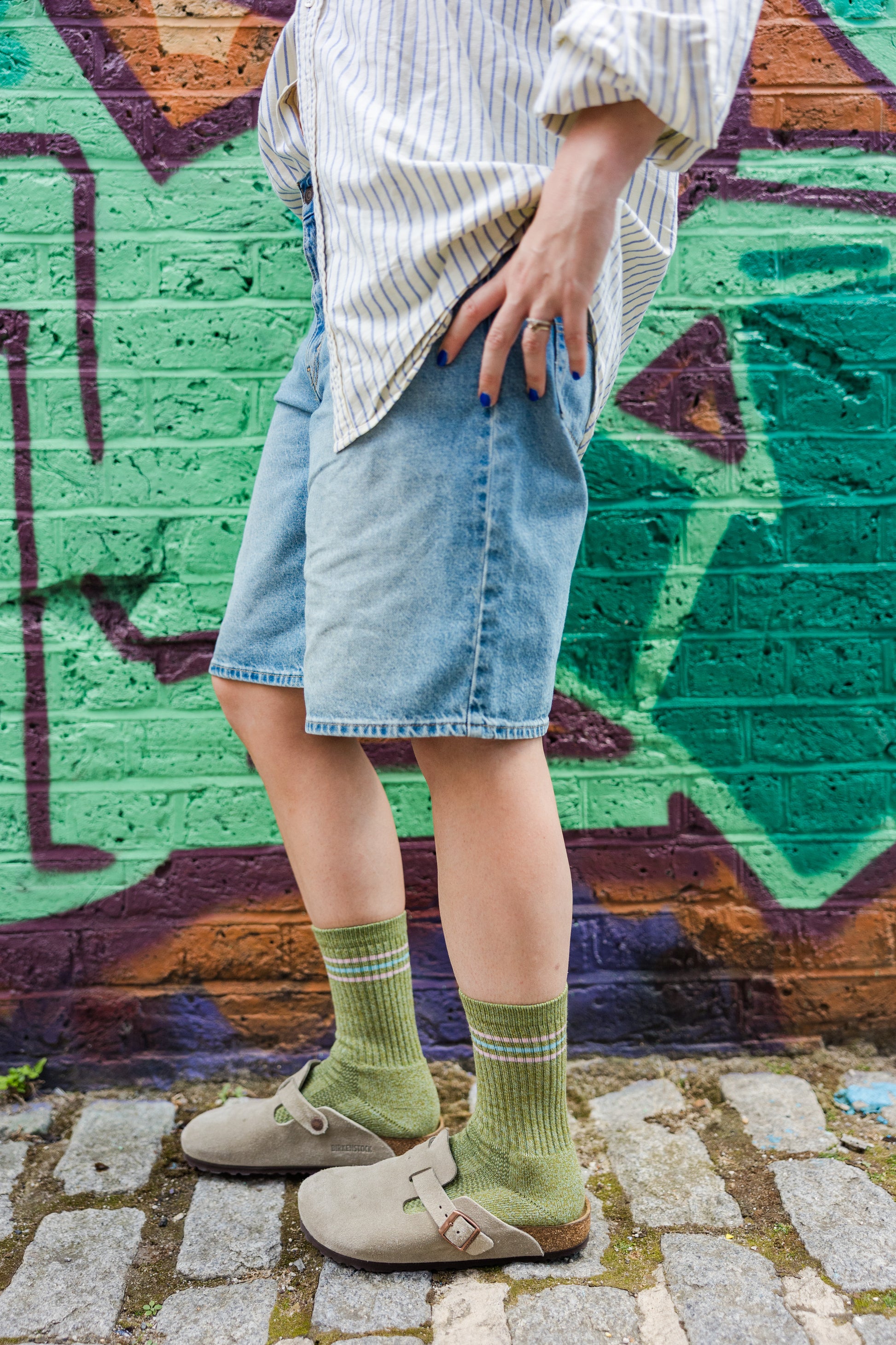 Person wearing denim levis bermuda shorts, striped shirt, and green socks standing in front of a graffiti-covered wall.

